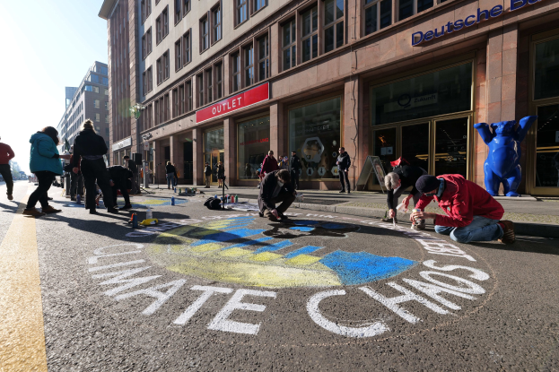 Eine Gruppe von Menschen sitzt vor einem Gebäude während einer Klimawandel-Demonstration in Berlin, umgeben von Flaschen und anderen Gegenständen, mit Bäumen und einem klaren blauen Himmel im Hintergrund.