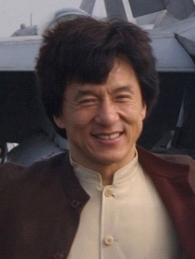 Jackie Chan steht strahlend vor einem Flugzeug unter einem klaren blauen Himmel, trägt ein weißes Hemd und eine schwarze Jacke mit ordentlich frisiertem Haar.