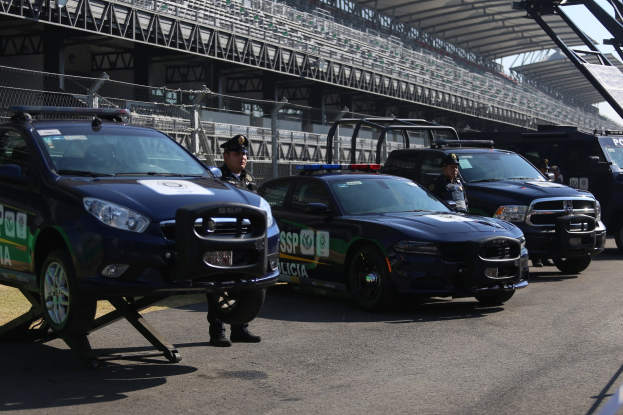 Polizeiautos vor einer Rennstrecke mit uniformierten Personen, Stadion und Absperrung im Hintergrund.