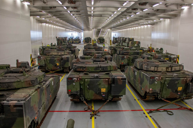 Ein grosser Militaraerraum mit zahlreichen Tanks in einer ordentlichen Reihe, beleuchtet von Oberlichtern und von Waenden umgeben.