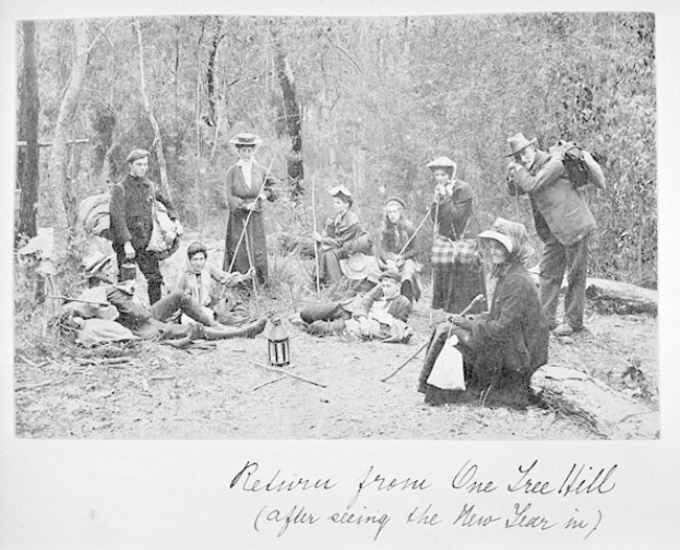Eine Gruppe von Menschen sitzt um ein Lagerfeuer im Wald. Einige tragen Hüte und Taschen, andere halten Stöcke. In der Mitte steht eine Laterne und im Hintergrund sind Bäume zu sehen.