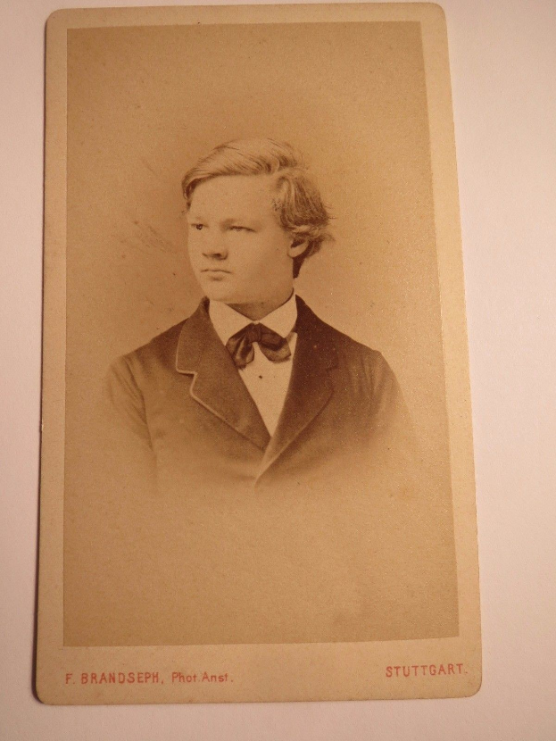 Schwarz-weiß-Foto eines jungen Mannes in Anzug und Fliege, identifiziert als F. Brandseph Stuttgart, aufgenommen in den frühen 1900er Jahren, befestigt an einer weißen Oberfläche mit Text unten.