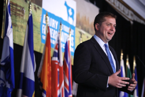 Ein Mann in Anzug und Krawatte spricht an einem Podium mit Mikrofon, umgeben von Flaggen und einem Bildschirm, wahrscheinlich zu einer Regierungszahlung an die Milchindustrie.