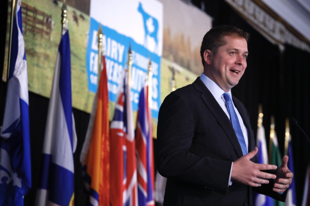 Ein Mann in Anzug und Krawatte spricht an einem Podium mit Mikrofon, umgeben von Flaggen und einem Bildschirm, wahrscheinlich zu einer Regierungszahlung an die Milchindustrie.