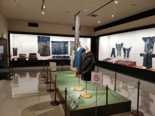 Ein Raum voller Schaufensterpuppen in traditioneller japanischer Kleidung, Vitrinen mit verschiedenen Kleidungsstücken, Informationsschilder, Stangen mit Ketten und Deckenleuchten.