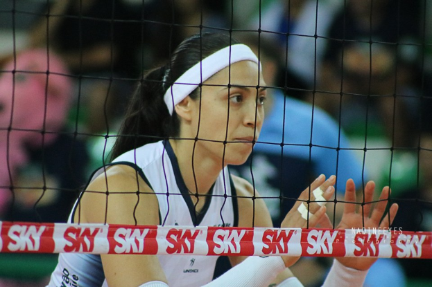 Frau im Volleyballspiel in Aktion auf einem Feld, mit einem Netz im Vordergrund und einem unscharfen Hintergrund, mit einem entschlossenen Gesichtsausdruck.