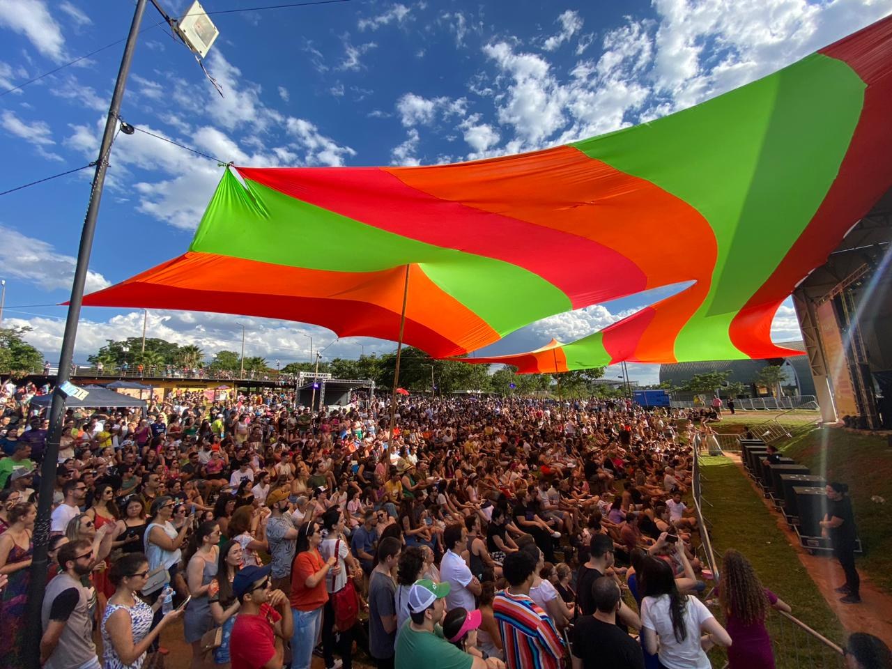 Eine große Menge steht vor einer Regenbogenflagge mit einem Gebäude, Bühnenlautsprechern, Bäumen und Wolken im Hintergrund während einer Festival-Jubiläumsfeier.