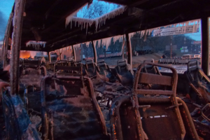 Ein zerstörter Bus mit verkohlten Sitzen und Eiszapfen am Dach, vor einer Kulisse aus Bäumen, Gebäuden und einem klaren Himmel.