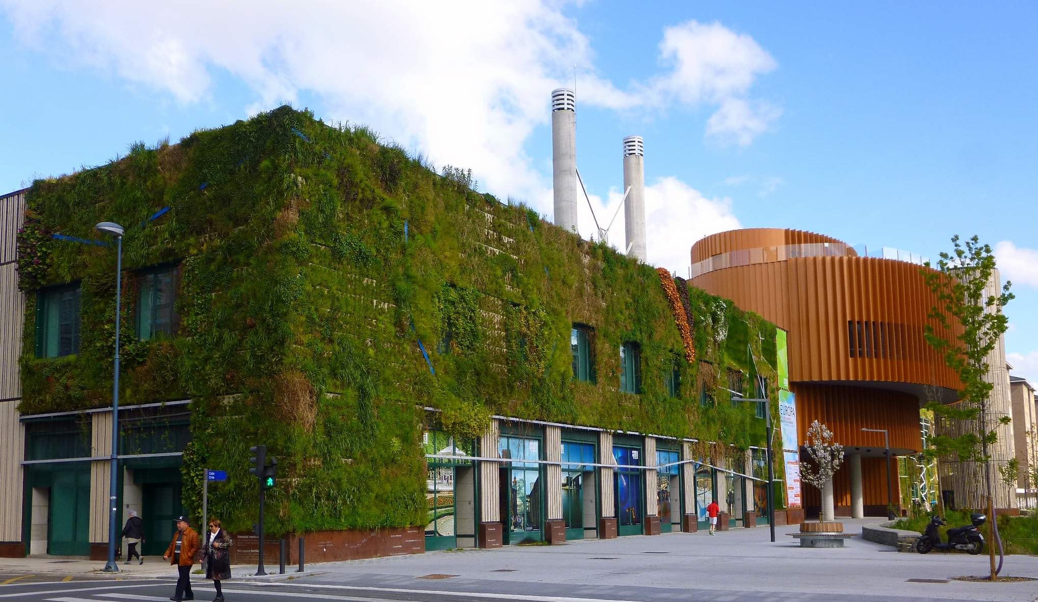 Ein großes Gebäude mit grüner Wand, umgeben von Straßeninfrastruktur, Kraftfahrzeugen, Fußgängern, Bäumen und einem bewölkten Himmel, Teil eines nachhaltigen Green Building-Projekts.