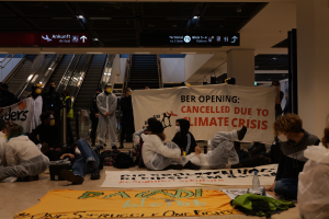 Eine Gruppe von Menschen sitzt auf dem Boden eines Flughafens und hält ein Banner in der Hand, auf dem "Berlin wegen Klimakrise gestrichen" steht. Um sie herum liegen Taschen, Flaschen und andere Gegenstände verstreut, im Hintergrund sind Rolltreppen, Schilder mit Text und Deckenleuchten zu sehen.