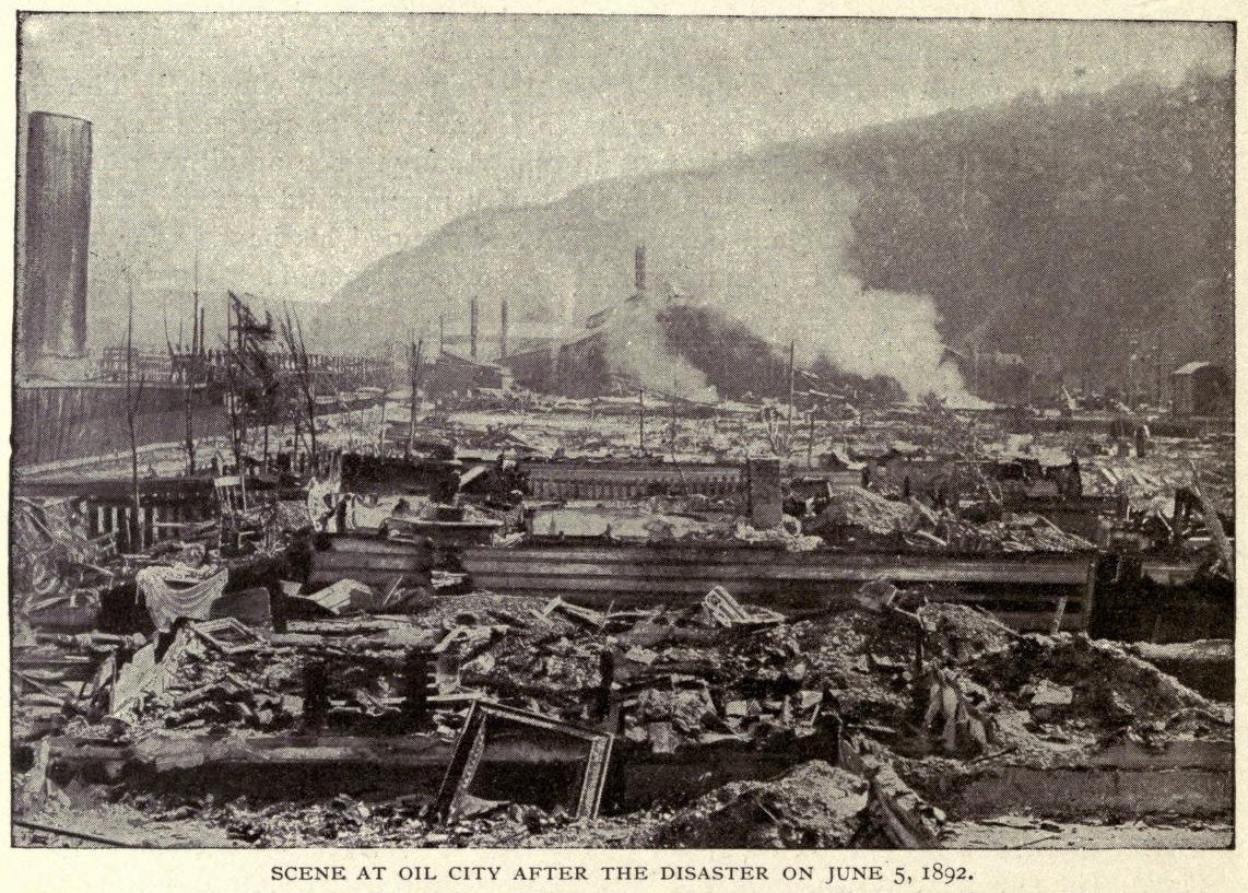 Schwarzes und weißes Foto einer Stadt nach einer Katastrophe am 5. Juni 1892, das beschädigte Gebäude, Strommasten, Rauch und einen Hügel im Hintergrund zeigt, mit Text am unteren Rand.