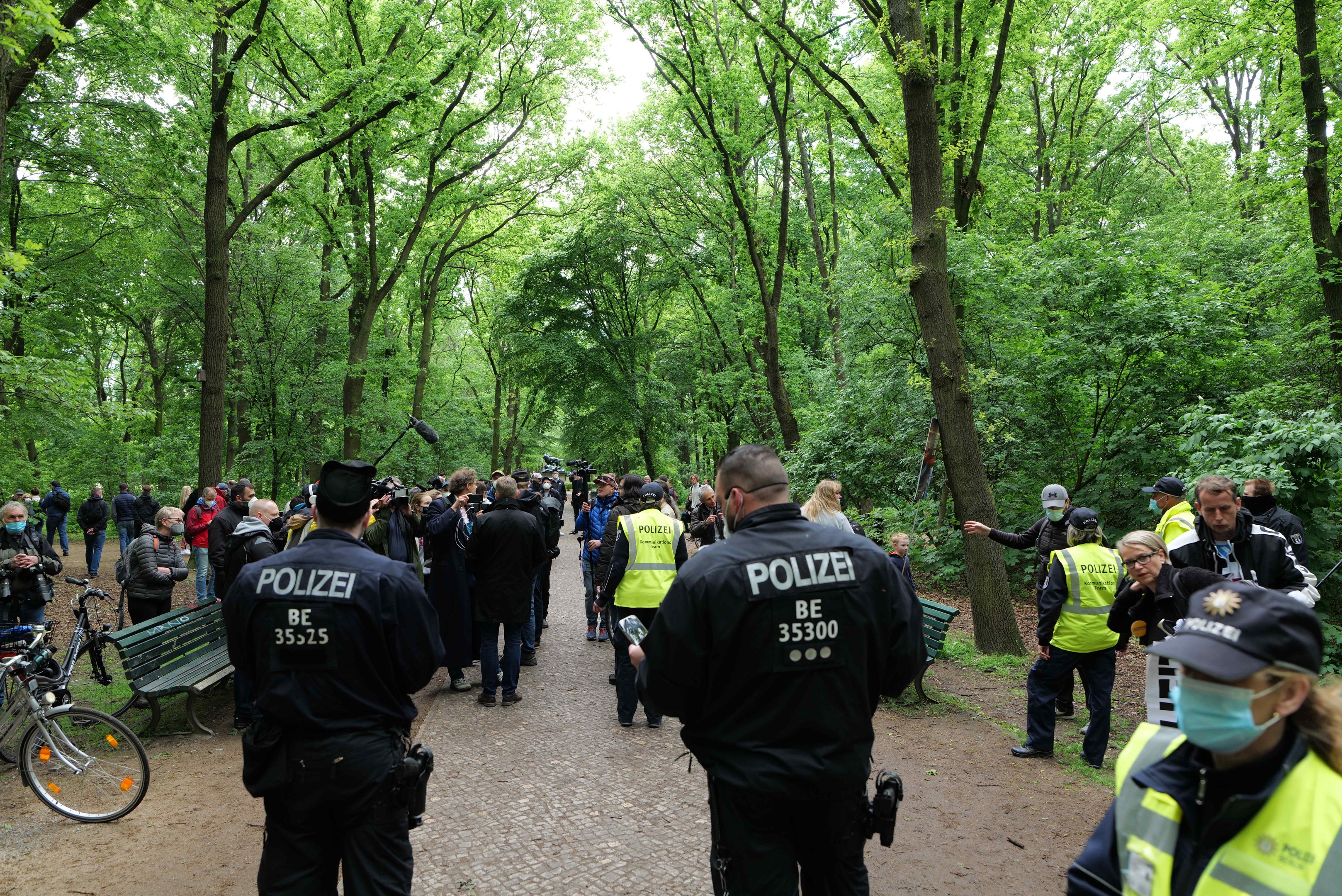 Polizeibeamte vor einer Menge während einer Outdoor-Veranstaltung, mit Fahrrädern und einer Bank im Vordergrund und Bäumen im Hintergrund.