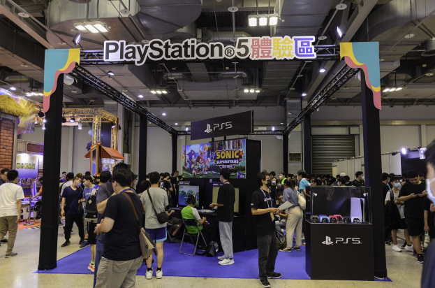 Eine Gruppe von Menschen an einem PlayStation-Stand während der Tokyo Games Expo, einige tragen Taschen oder sitzen auf Stühlen, mit beleuchteten Texttafeln, Deckenbeleuchtung, einem Bildschirm im Hintergrund und einem Tisch mit Gegenständen rechts.