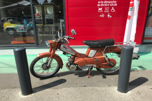 Ein oranges Mofa steht auf der Straße vor einem roten Gebäude mit Glaswänden, mit zwei schwarzen Pfosten im Vordergrund und einer Flagge mit Text an der Wand rechts.