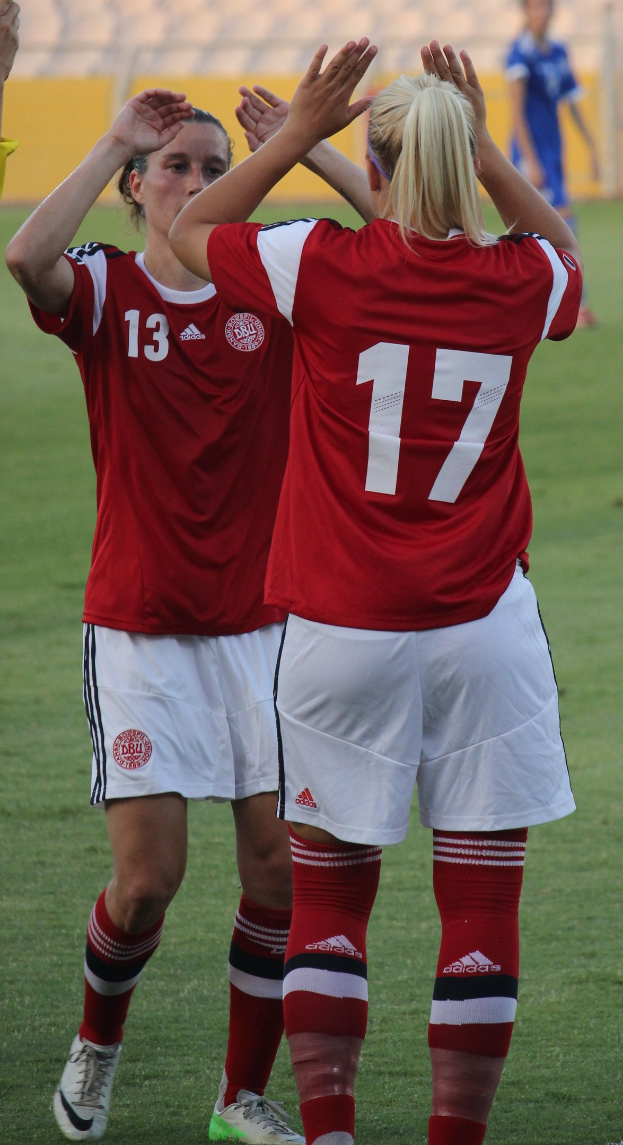 Zwei Frauen in roten T-Shirts und weißen Shorts klatschen auf einem Fußballfeld mit einer Person in einem blauen Kleid und einem gelben Schild im Hintergrund.