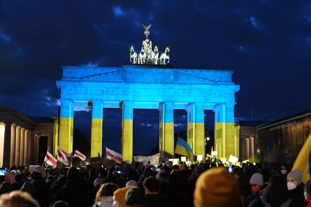 Eine Menschenmenge steht vor dem Brandenburger Tor in Berlin, Deutschland, mit Fahnen und Plakaten in den Händen, mit einer Banner auf der rechten Seite und dem Tor mit Statuen und Säulen geschmückt, unter einem bewölkten Himmel.