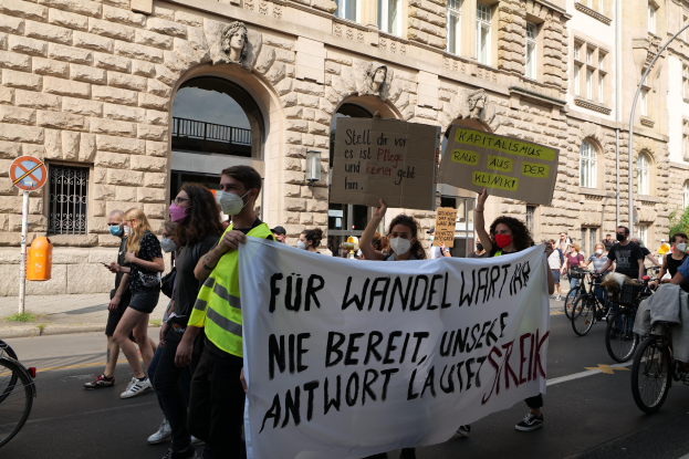 Eine Gruppe von Menschen marschiert bei einer Protestaktion gegen die Anti-Kriegs-Bewegung in Berlin, einige halten Schilder und Banner, andere fahren Fahrräder, im Hintergrund ein Gebäude mit Fenstern, Bögen, Säulen und Skulpturen.