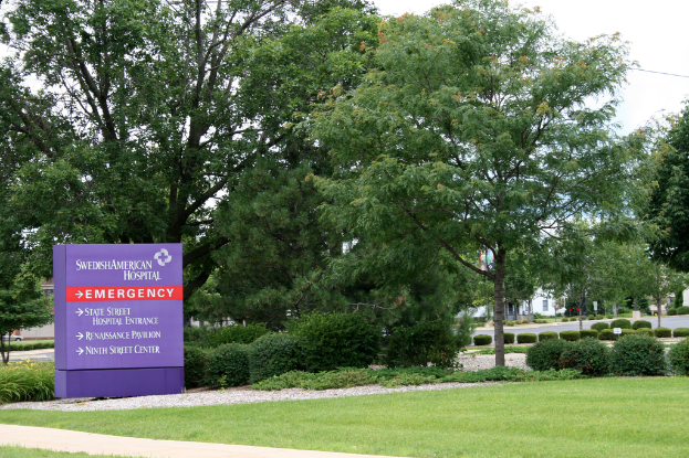 Ein grasbewachsener Bereich mit einem violetten Notfallschild, auf dem "NOTFALL" in fetter Schrift steht, umgeben von Bäumen und Sträuchern, mit Gebäuden und einer Straße im Hintergrund.