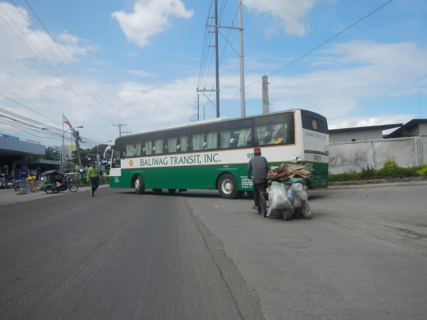 Ein grüner und weißer Bus fährt auf einer Straße mit Gebäuden, Fußgängern, einem Radfahrer, Strommasten, Bäumen und einem bewölkten Himmel.