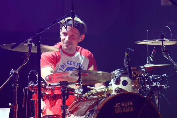 Ein Mann in einem roten und weißen T-Shirt spielt Schlagzeug auf der Bühne während eines Konzerts und lächelt umgeben von Mikrofonen.