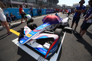Ein Formula-E-Auto mit weißer, blauer, roter und schwarzer Lackierung steht auf einer Rennstrecke, umgeben von Menschen, mit einem Schirm im Vordergrund und Rennstrecken-Infrastruktur im Hintergrund unter einem klaren blauen Himmel.
