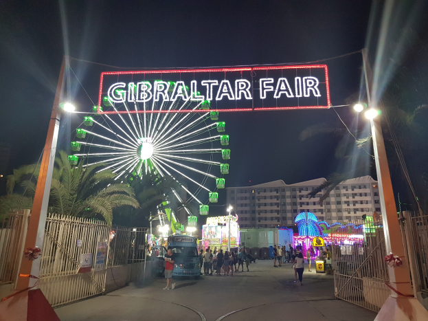Eingang zur Gibraltar-Messe nachts mit beleuchtetem Riesenrad, Menschenmenge, Leuchtschilder, Bäumen und Gebäuden im Hintergrund.