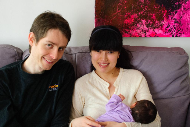 Eine Frau in einem wei├čen Hemd h├Ąlt ein Baby und l├Ąchelt, waehrend sie auf einem Sofa sitzt, neben einem Mann in einem schwarzen T-Shirt, der ebenfalls l├Ąchelt, mit einem rosafarbenen Fotoраhmen an der wei├čen Wand dahinter.