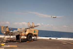 Ein militärisches Fahrzeug ist auf dem Deck eines Flugzeugträgers positioniert, mit einem Drohnenflug darüber; die Szene ist gegen einen bewölkten Himmel und sichtbares Wasser im Hintergrund gesetzt.