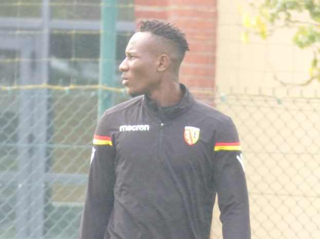 Ein Mann in einer schwarzen Jacke steht auf einem Fussballfeld, umgeben von einem Zaun und einem Baum, und sieht konzentriert aus während eines Trainingssitzungs.