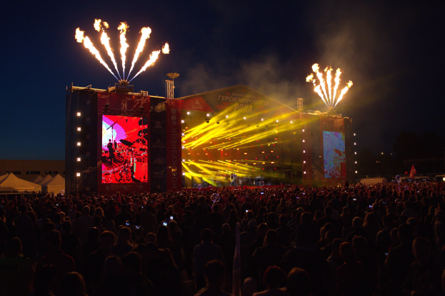 Eine große Menschenmenge bei einem Musikfestival schaut auf der hell erleuchteten Bühne Performern zu, mit Zelten links und Feuerwerk am dunklen Himmel dahinter.
