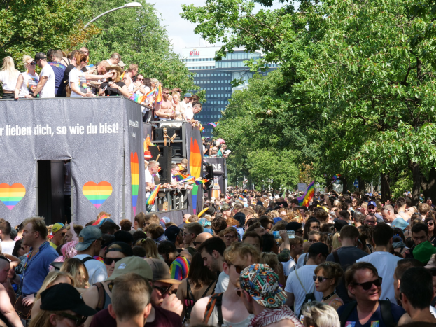 Eine große Menge steht vor einem Lastwagen bei der Christopher Street Day Parade in Berlin, viele tragen Mützen und Schutzbrillen, einige halten Fahnen, mit einem Banner auf dem Lastwagen und Bäumen, Gebäuden und einem Laternenpfahl im Hintergrund unter einem bewölkten Himmel.