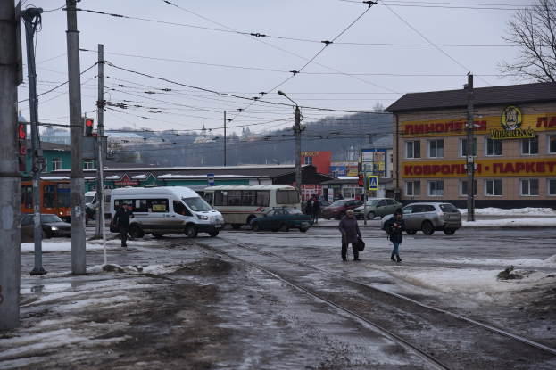 Eine Gruppe von Menschen geht eine Straße neben einer Bahnschiene mit Fahrzeugen, Strommasten, Ampeln, Gebäuden, Bäumen und einem Van im Hintergrund.