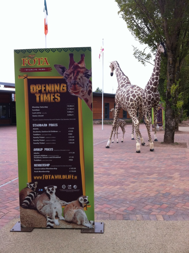 Giraffen stehen neben Bäumen, Schuppen und Flaggen, mit einer Tafel, die Text, Logos und Tierbilder zeigt.