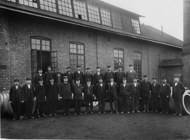 Eine Gruppe von Menschen in schwarzen Anzügen und Mützen vor einem Gebäude mit sichtbaren Türen und Fenstern.