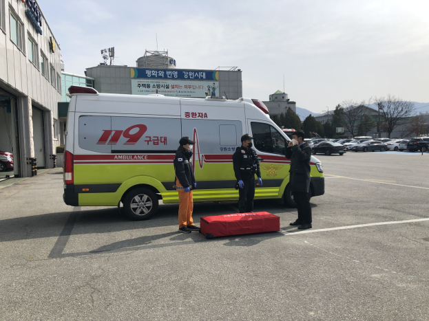 Ambulanz auf einem Parkplatz neben einem Gebäude mit drei Personen auf der Straße davor geparkt, links ein Gebäude mit Glasfenstern, andere Fahrzeuge, Bäume, Berge und Himmel im Hintergrund.