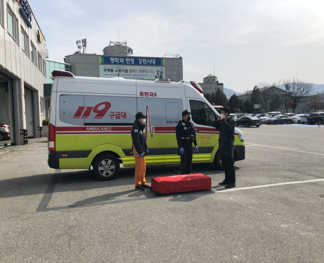 Ambulanz auf einem Parkplatz neben einem Gebäude mit drei Personen auf der Straße davor geparkt, links ein Gebäude mit Glasfenstern, andere Fahrzeuge, Bäume, Berge und Himmel im Hintergrund.