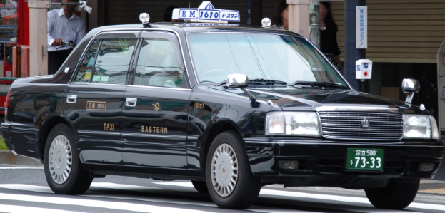 Ein schwarzes Taxi fährt die Straße entlang, mit einem Radfahrer im Vordergrund, Gebäude, Masten und Schilder im Hintergrund.