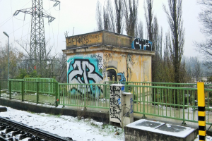 Ein Bahngeleis mit Graffiti an der Seite, umgeben von Gelöndern, Pfosten, Lichtern und Bäumen, mit Törmen und Dröhnen im Hintergrund und Schnee am Boden.