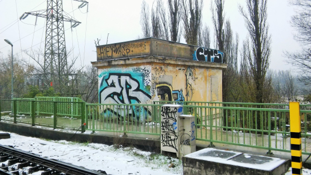 Ein Bahngeleis mit Graffiti an der Seite, umgeben von Gelöndern, Pfosten, Lichtern und Bäumen, mit Törmen und Dröhnen im Hintergrund und Schnee am Boden.