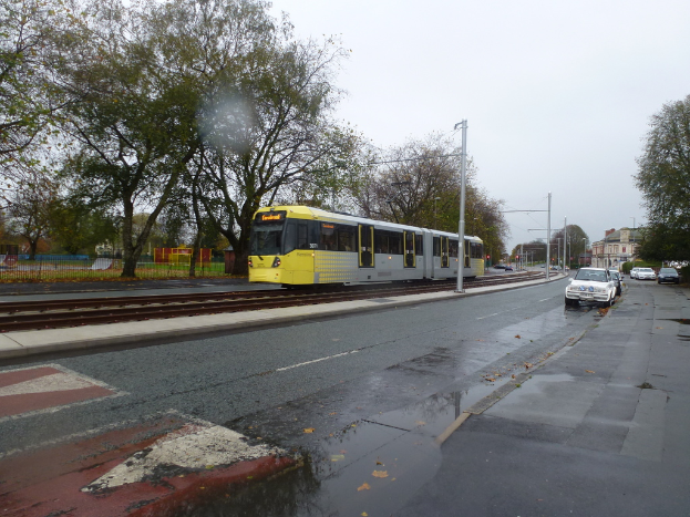 Eine belebte Straßenszene mit Fahrzeugen, Gebäuden, Polen, einer Bahnstrecke mit einem Zug, Bäumen und einem Zaun unter einem klaren Himmel.