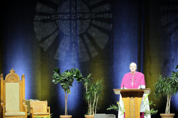 Ein Mann in einem rosa und weißen Kleid steht neben einem Rednerpult in einem Auditorium, mit Pflanzen und zwei Stühlen daneben und einer bunten Wand im Hintergrund.