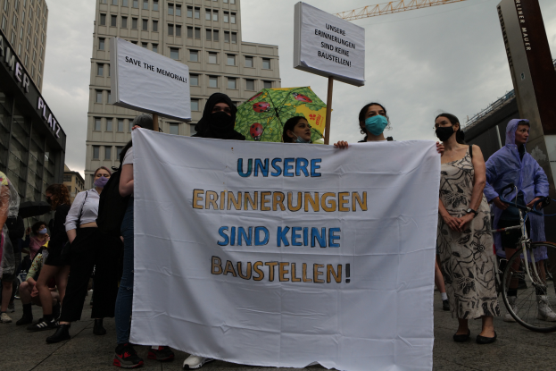 Eine Gruppe maskierter Personen mit einer Tafel mit der Aufschrift 'Unsere Erinnerungen sind keine Baustellen' steht vor einem Gebäude, wobei eine Person ein Fahrrad und eine andere einen Schirm hält, sowie Tafeln mit Text, während im Hintergrund Gebäude, ein Kran und ein bewölkter Himmel zu sehen sind.
