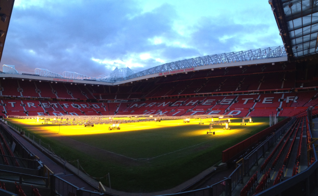 Old Trafford Stadium, Heimat von Manchester United, mit einem grünen Feld, umgeben von Sitzplätzen, hellen Lichtern, einer schuppenähnlichen Struktur auf der rechten Seite und einem Himmel mit weißen Wolken.