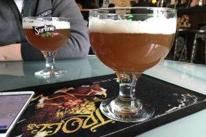 Zwei Gläser goldenes Bier auf einem Tisch neben einem Handy, mit Menschen und gerahmten Wandbildern im Hintergrund.