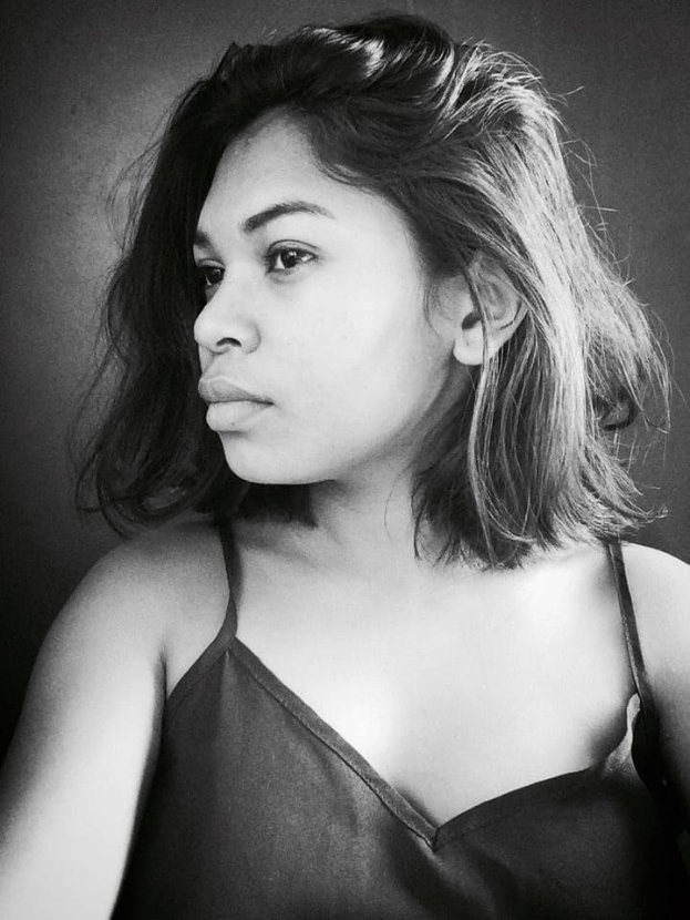 Eine selbstbewusste Frau in einem schwarzen Kleid posiert in einem Schwarz-Weiß-Foto, steht mit selbstbewusster Haltung vor einer Wand, ihr glatter Bob und dezentes Make-up unterstreichen ihre natürliche Eleganz.