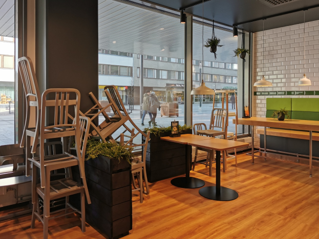 Ein Restaurant in Berlin mit Holztischen und -stühlen, die durch ein großes Fenster zu sehen sind, umgeben von Pflanzen und Beleuchtung, mit Fußgängern und Gebäuden draußen.