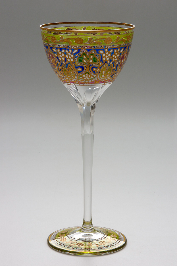Ein Weinglas mit grün-goldener Verzierung steht auf einer weißen Oberfläche und zeigt einen Stiel und eine schalenförmige Form mit breiter Basis und engem Hals.