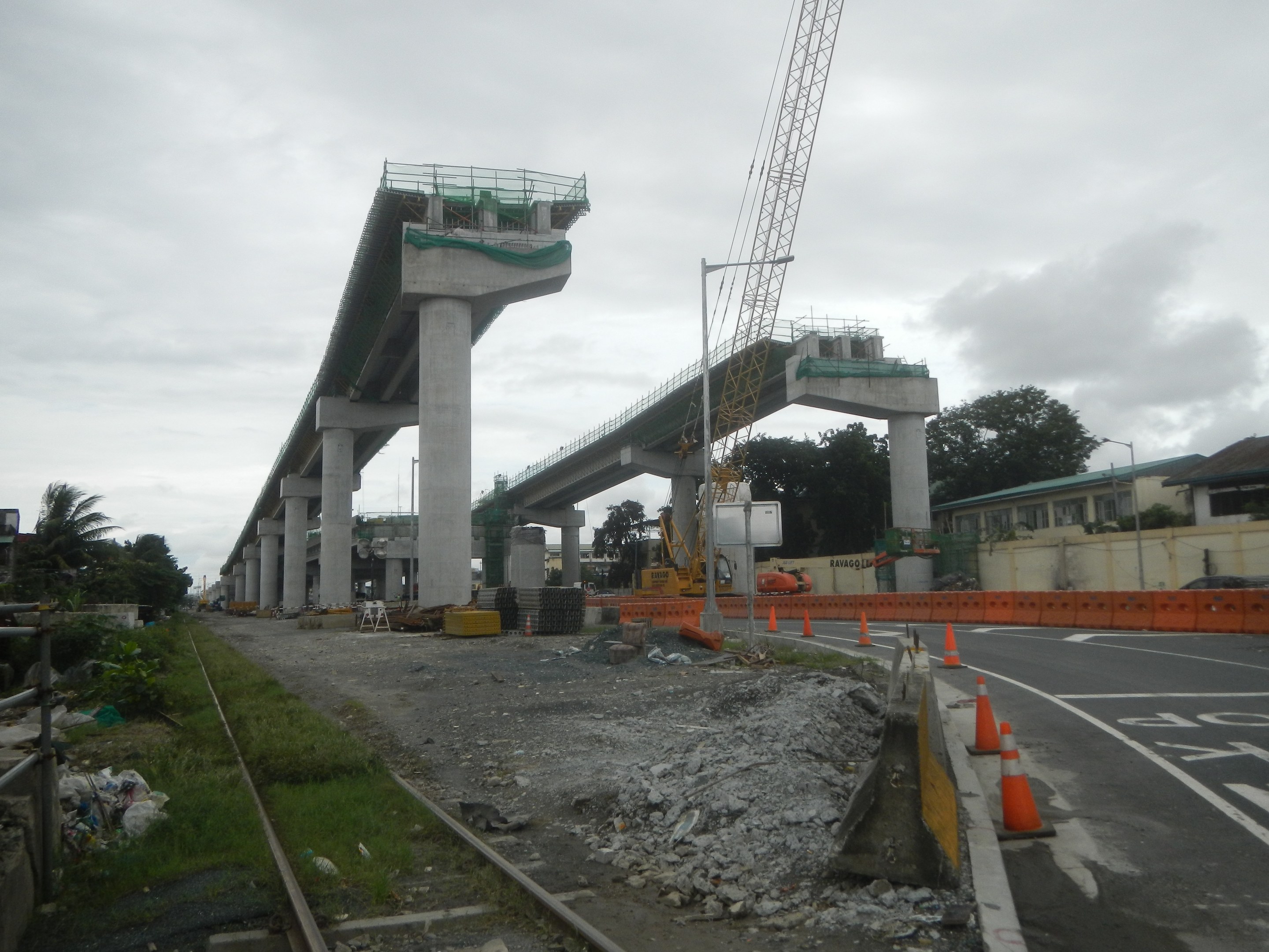 Baustelle mit einer Brücke im Hintergrund, Straße mit Verkehrskegeln markiert, Bahnschiene auf der linken Seite, verstreute Steine und Gras, Bäume und Gebäude auf beiden Seiten und ein bewölkter Himmel.