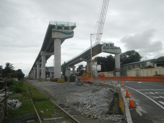 Baustelle mit einer Brücke im Hintergrund, Straße mit Verkehrskegeln markiert, Bahnschiene auf der linken Seite, verstreute Steine und Gras, Bäume und Gebäude auf beiden Seiten und ein bewölkter Himmel.