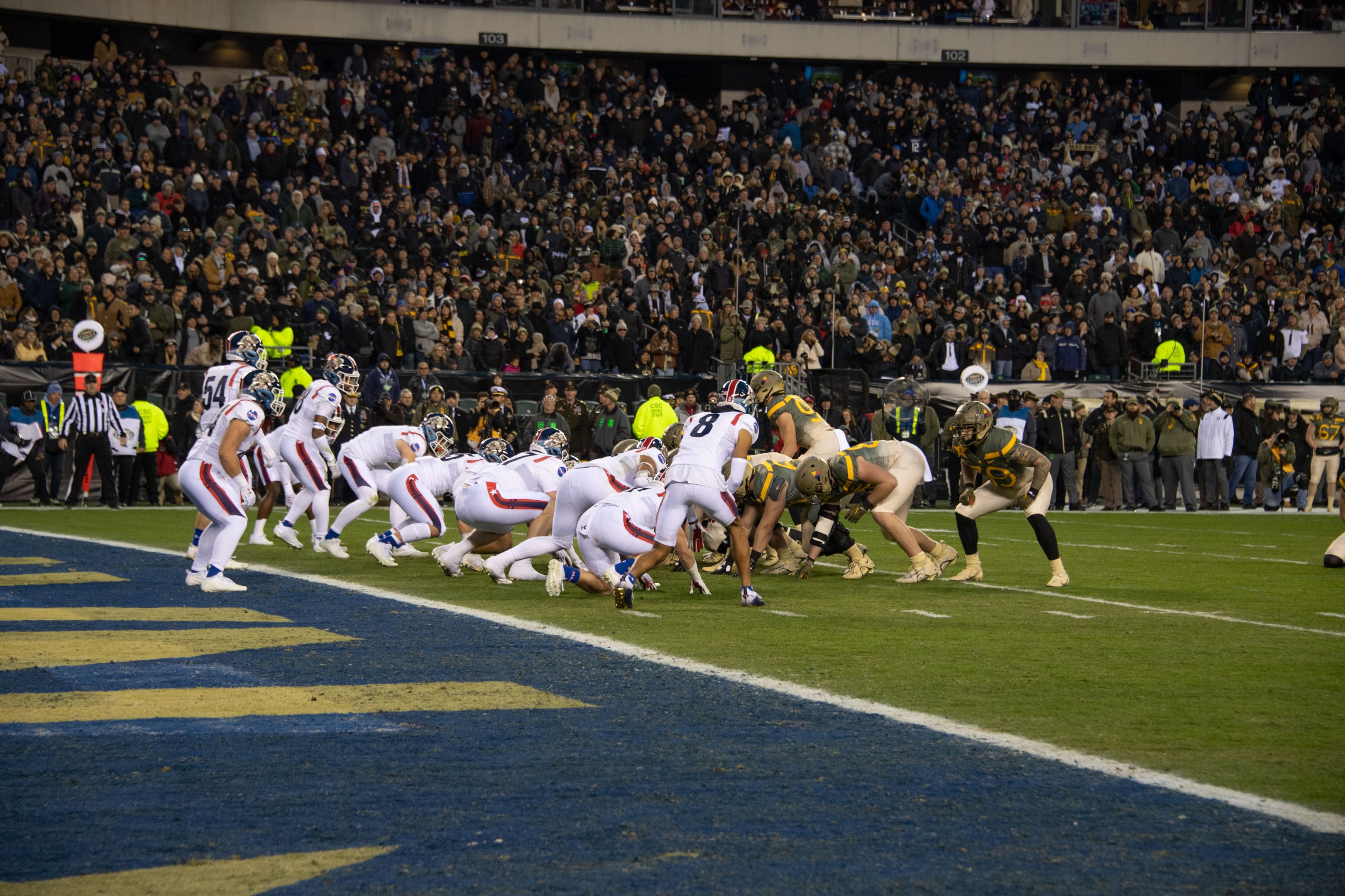 Ein Fussballspiel im Gange mit Spielern in Sportkleidung und Helmen auf dem Feld, Zuschauern, Gel"andern und Tafeln mit Text im Hintergrund.